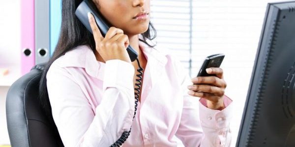 woman-at-desk-texting-while-listening-on-phone-600×600